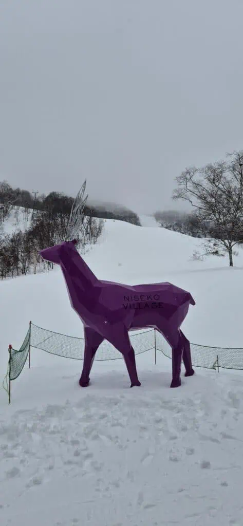 A purple deer statue with  Niseko Village written on it, set in front of the slopes