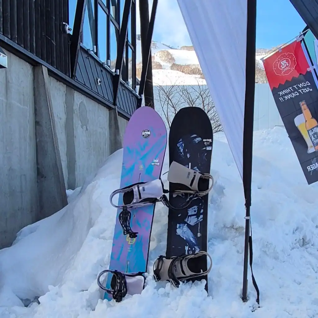 Two rental snowboards sit next to each other propped up in the snow.